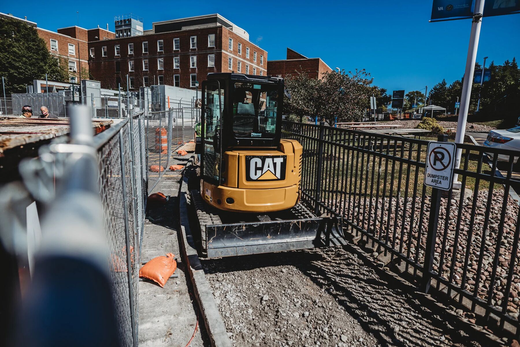 Street-level view of the fenced Syracuse VA work zone with campus buildings behind